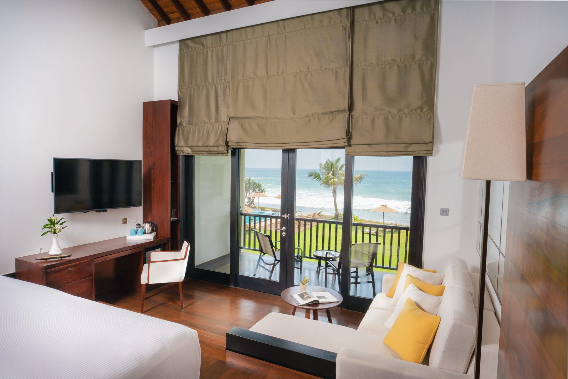 The sitting area of an Ocean Room: a white sofa with yellow cushions, a desk and a wall-mounted TV, with glass doors to a balcony over the lawn, the pool and the sea