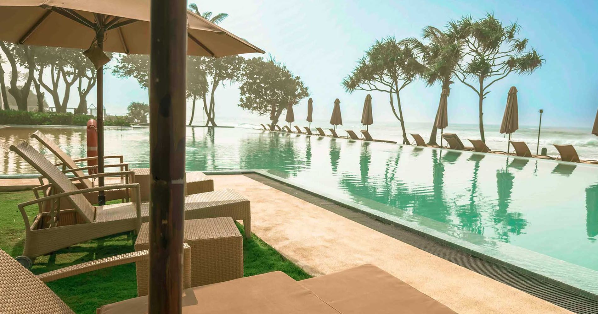 Wicker sun loungers and a parasol on the lawn beside the infinity pool, with loungers, furled parasols and trees along its far edge and the sea beyond
